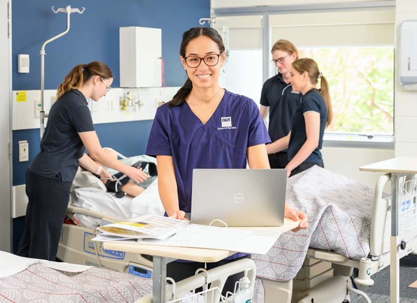 A woman in blue nursing scrubs stands between hospital beds. In front of her is a folder and a laptop, behind her three students are running a procedure a mannequin.