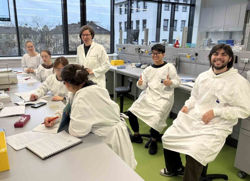 A group of male and female students wearing lab coats sit at benches in a laboratory. One of the male students is smiling at the camera and giving the thumbs up while the others work on a task.