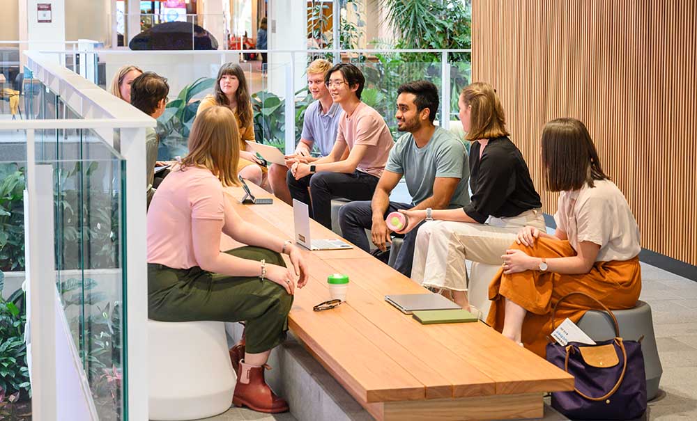 A group of students sit at a long table in a modern building. They are deep in discussion.