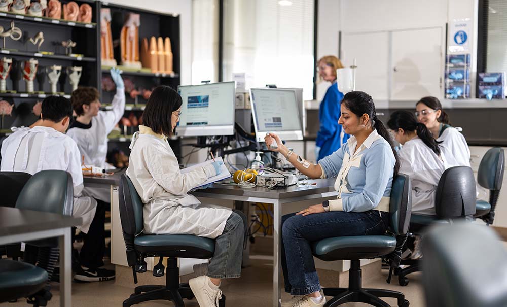 Groups of students sit at group desks in a classroom. There are bookshelves around the edges of the room featuring detailed models of different body parts.