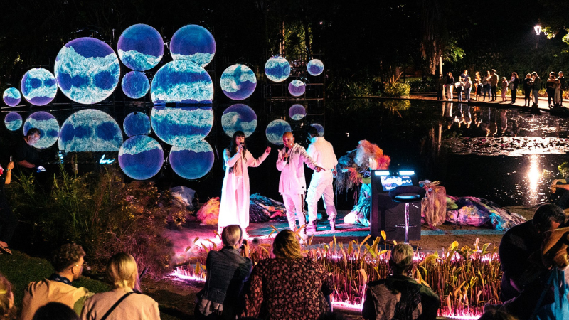 a group of singers perform in the dark in front of a lake with colour light projections of blue bubbles shown behind them