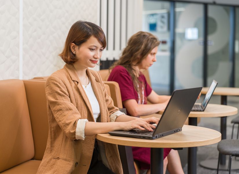 Two female postgraduate students working on their laptops in a bright space on campus