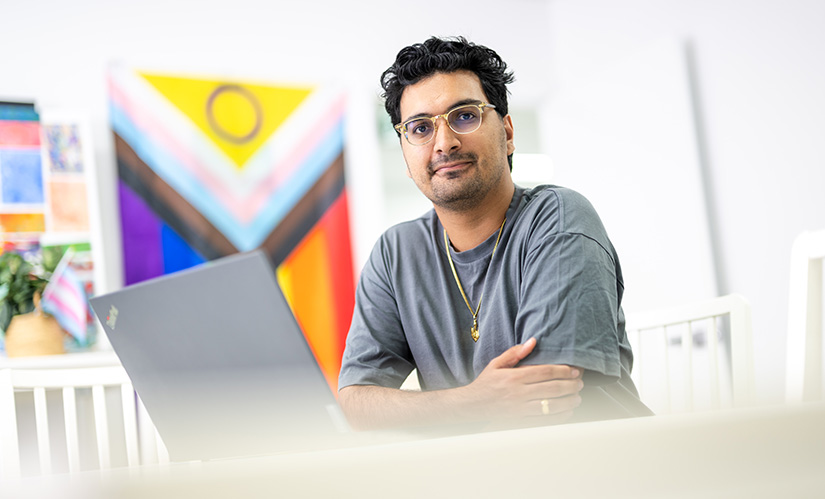 A man with a laptop sits at a table, behind him are LGBTQIA flags and posters.