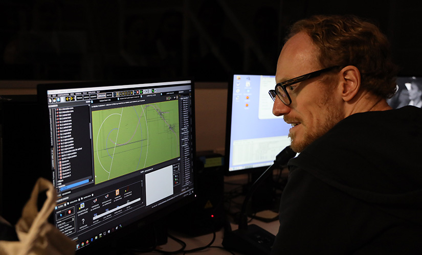 A male researcher sits at a console with screens and controls in front of him.