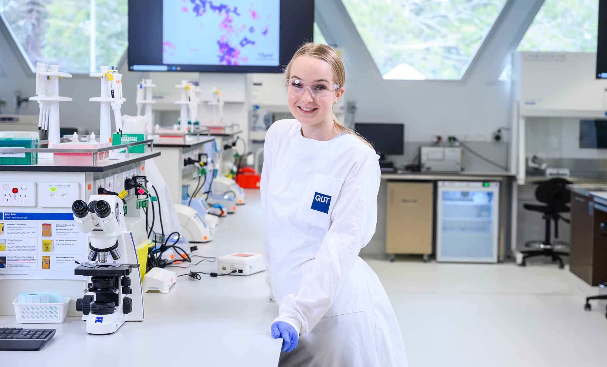 A female student in a lab coat stands in front of a microscope.