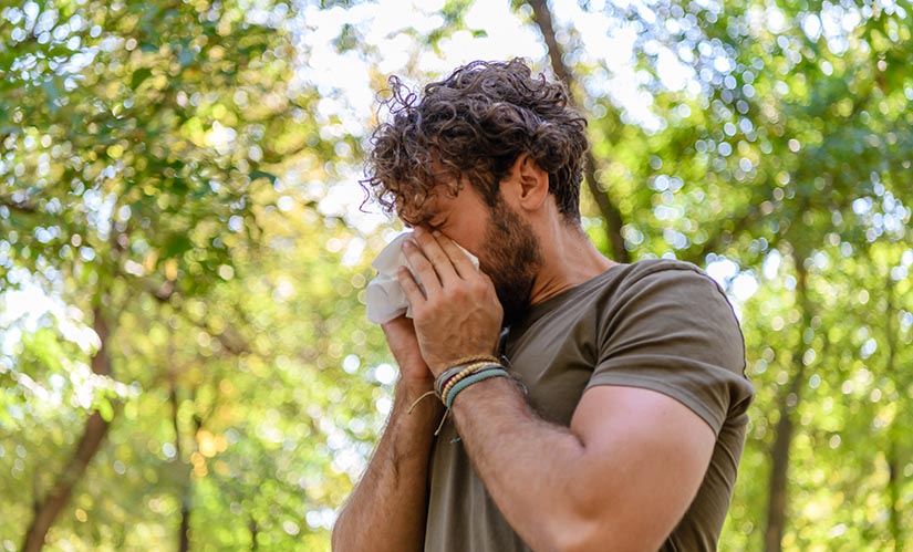 A man walking through a park sneezes into a tissue.