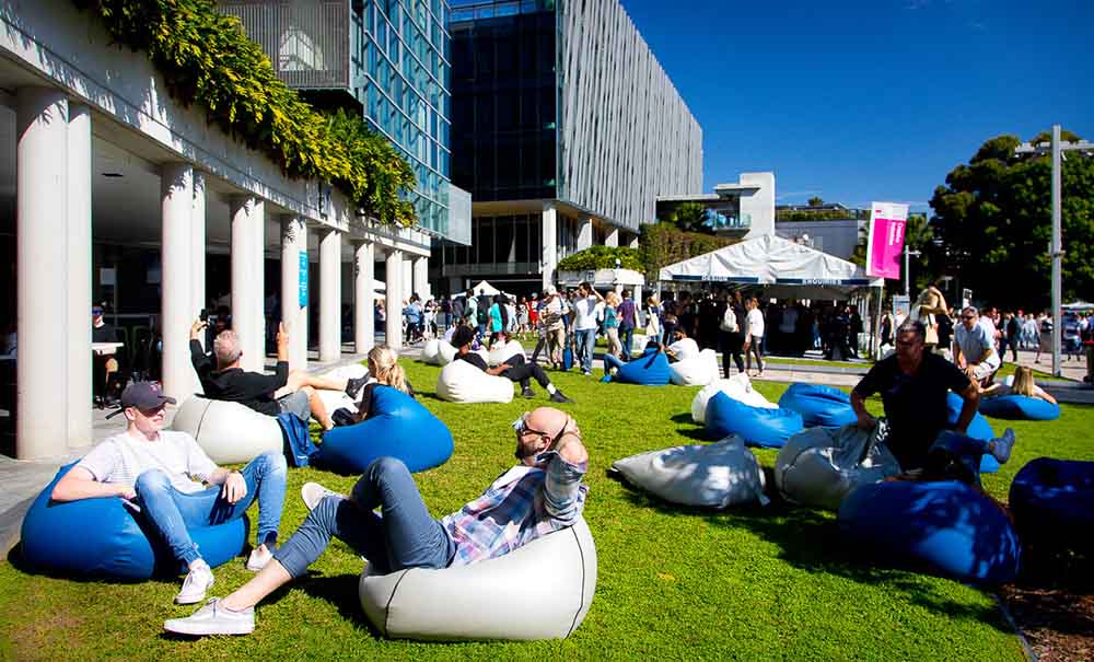 People attending QUT Open Day, resting on blue and white bean bags on a sunny lawn in front of tents, banners and modern buildings.