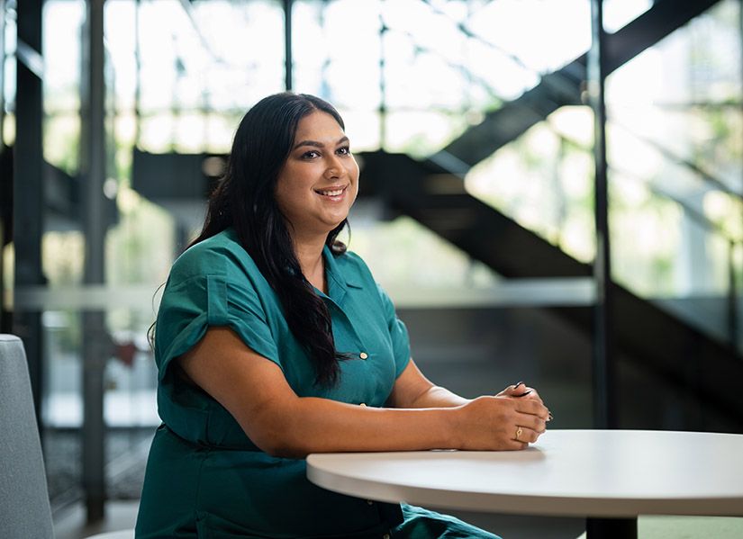 A woman in a green dress sits at a table in a modern office foyer. She is smiling at someone in the distance.