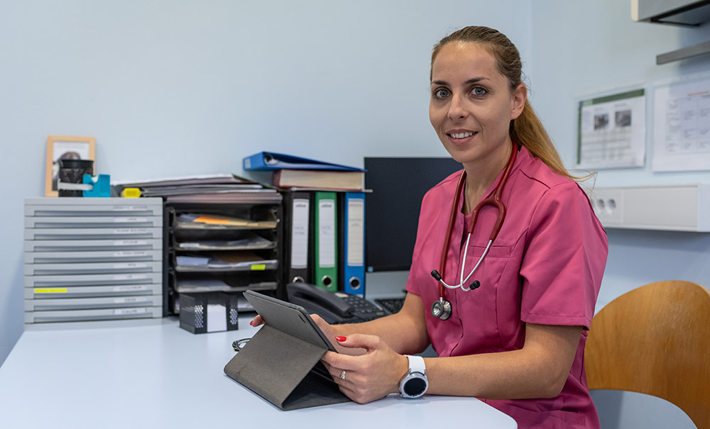 A female nurse practitioner in bink scrubs sits at a desk. Behind her are folders, a computer and a filing tray.