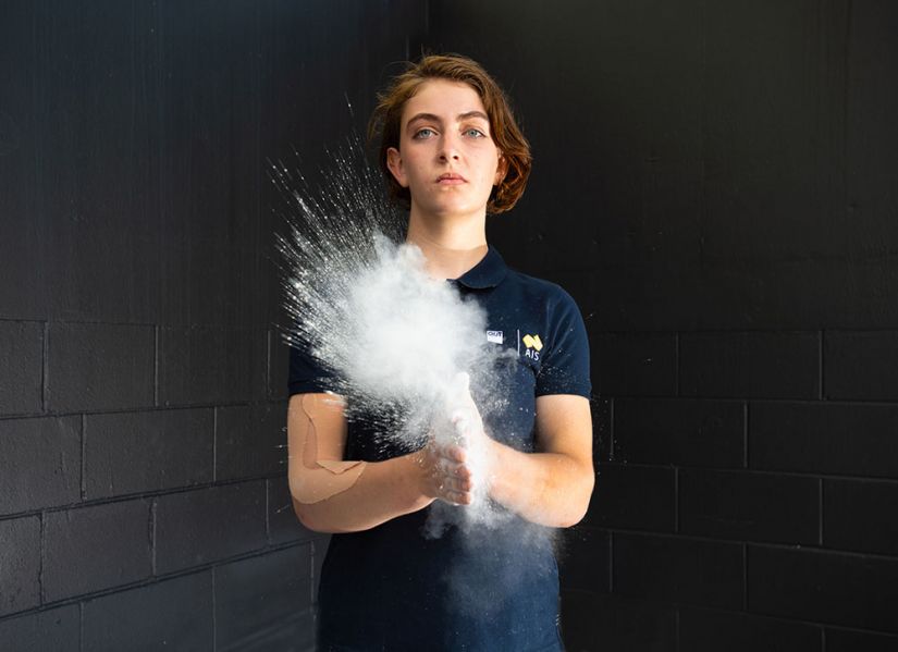 A woman in a sports uniform claps hands together releasing a cloud of chalk powder used to help with her grip.