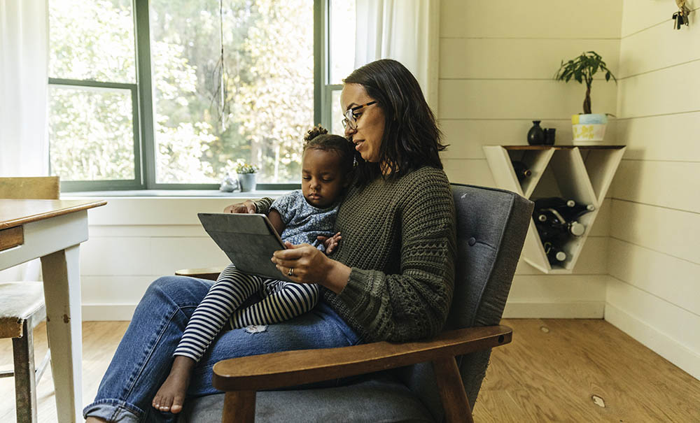 A mother in a country living room balances a small girl and an iPad on her lap as she logs in to a telehealth appointment. 