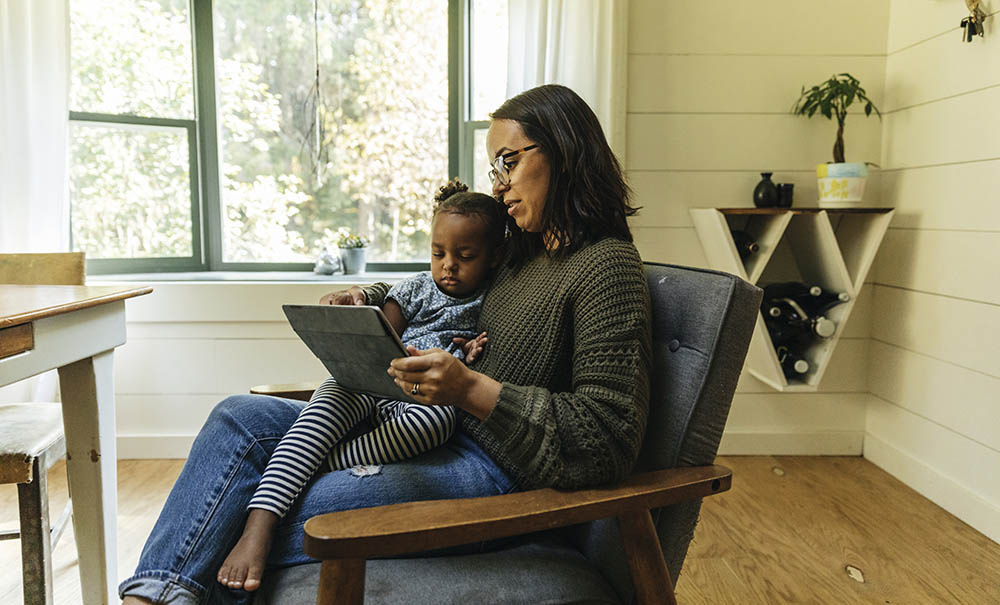 A mother in a country living room balances a small girl and an iPad on her lap as she logs in to a telehealth appointment.