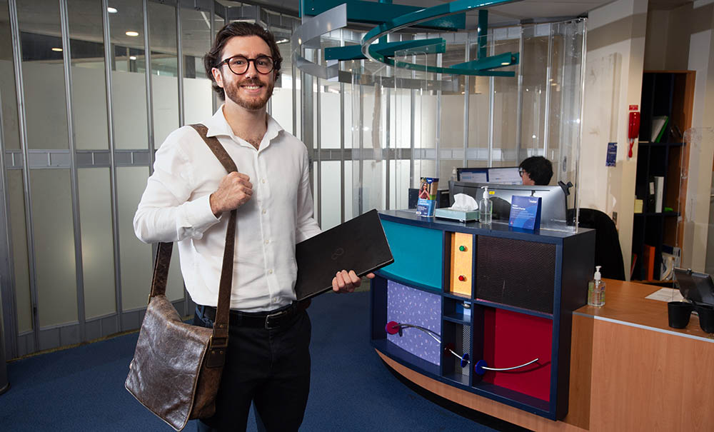 A man wearing a white collared shirt, carrying a satchel and a laptop, walks into a telehealth centre.