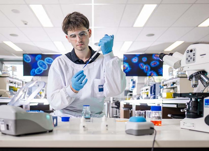 A male student in a white lab coat sits at a bench in a laboratory. He is using a pipette to transfer fluid into a vial.