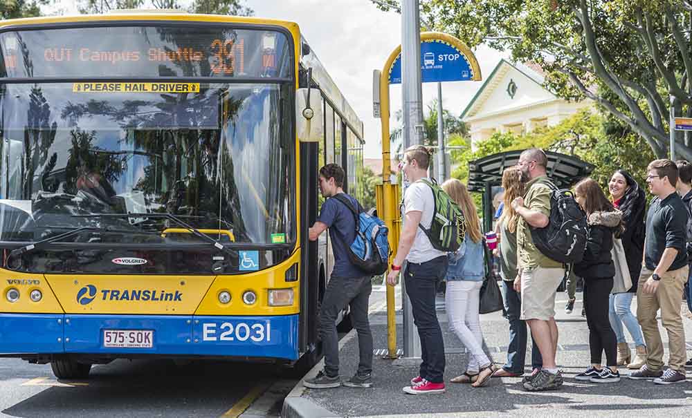 A group of students line up on the sidewalk to board the QUT intercampus shuttle bus.