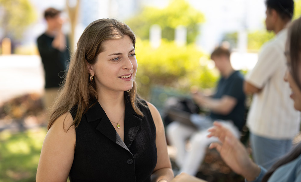 A woman stands in a leafy area outside, she is listening to the person standing in front of her.