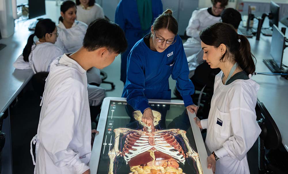 A teacher and two students stand over a table with a screen display built into it. On the screen is an anatomically correct image of a human showing the skeleton and the digestive system. The teacher is pointing out a feature on the display.