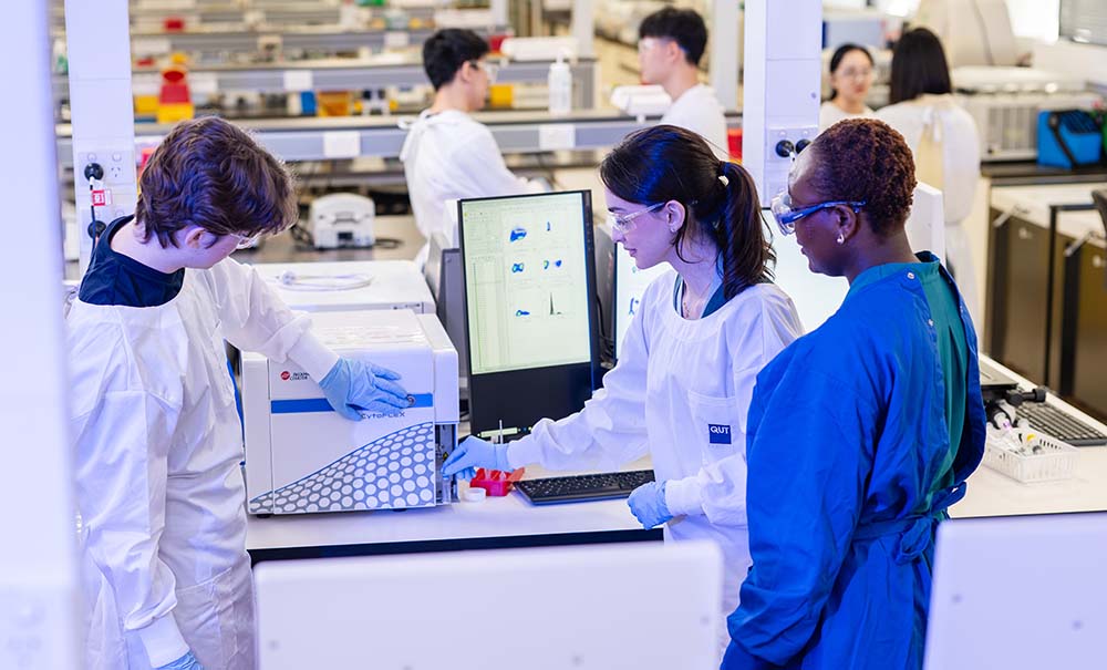 Two students and a teacher stand infront of a machine the size of a microwave. One of the students is reaching down to remove a vial from an opening at the front of the machine.