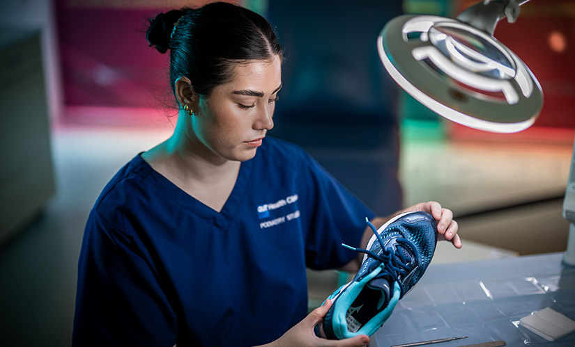 A female student in scrubs looks down at a sneaker she is holding in her hands.