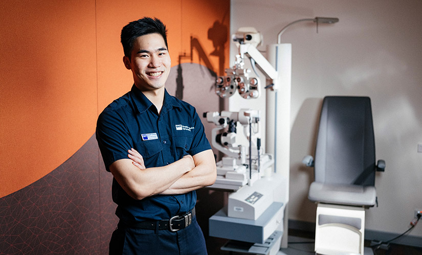 A male student stands in front of an eye exam chair.