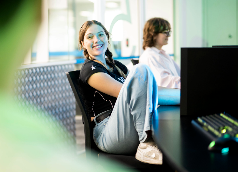 female games student sitting on a swivel chair with her leg pulled up on the seat casually, smiling towards her classmate in a computer lab on campus