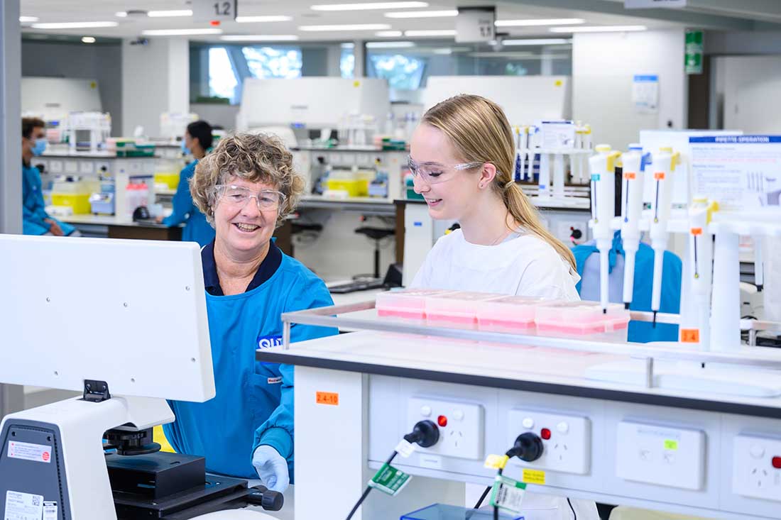 A student wearing a lab coat stands in front of a microscope while a teacher explains how to perform a test.