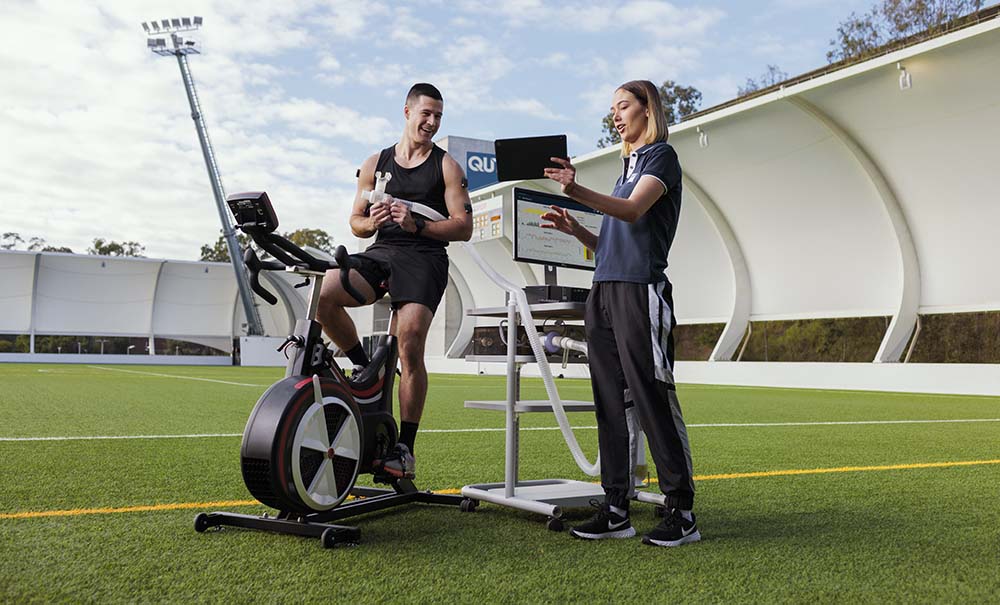 Kaine sits on an exercise bike holding equipment for measuring sports performance and listening to feedback from a colleague.