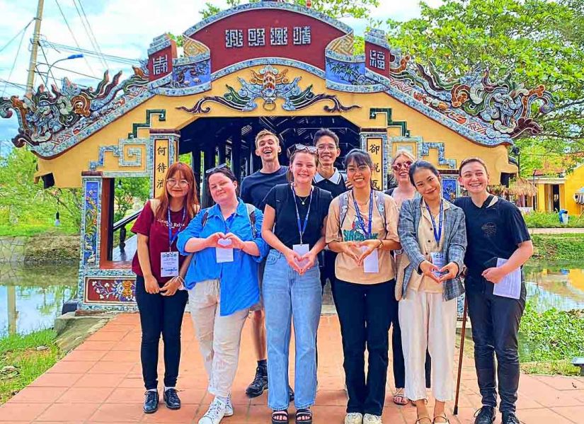 A group of smiling students stands in front of an ornate Vietnamese building.