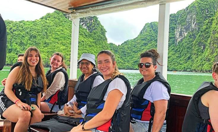 A group of students wearing life jackets sit in a boat on a lake. Behind them there are giant mountains that emerge directly out of the water.
