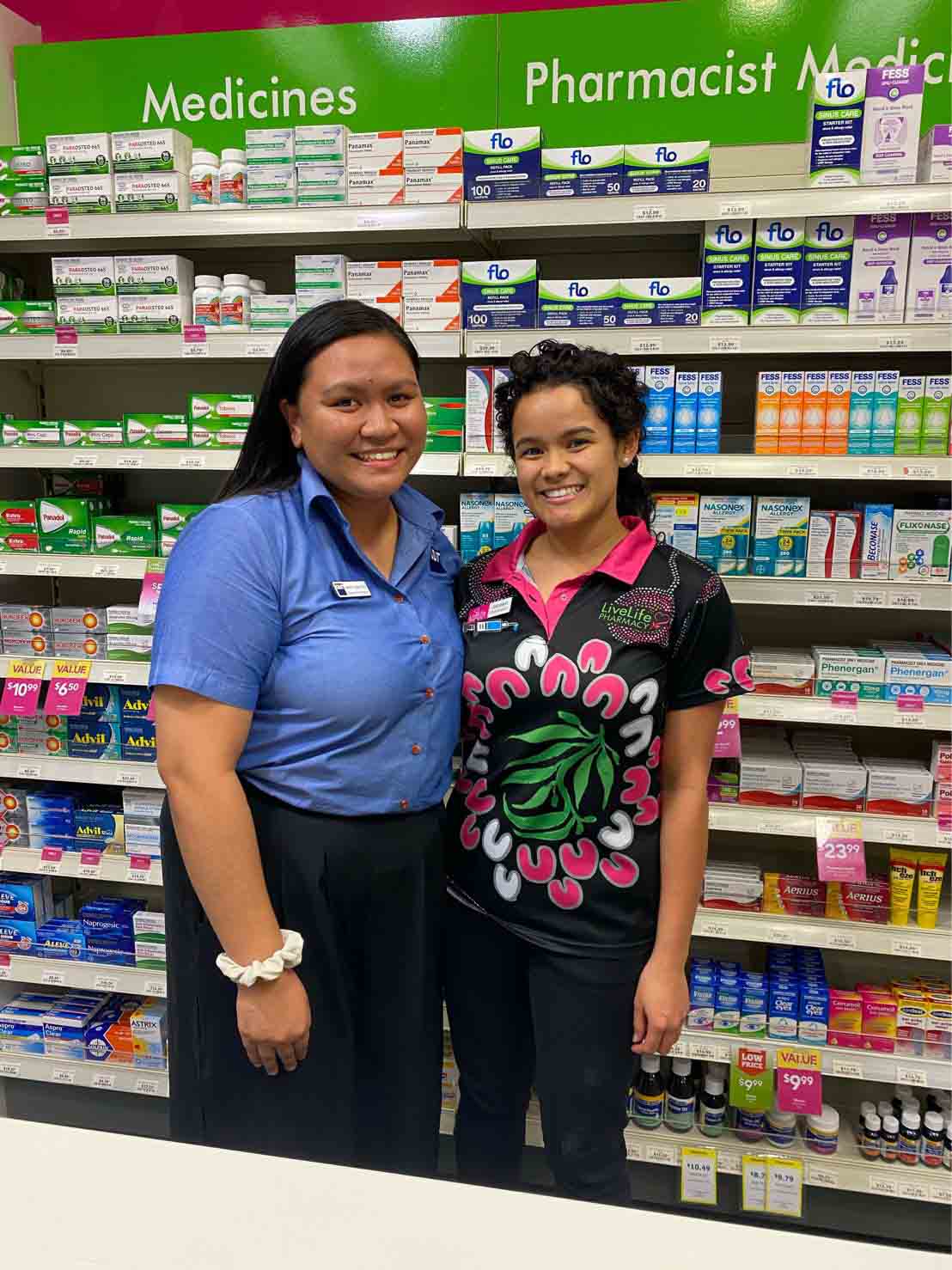 Two smiling women in pharmacy uniforms stand in front of a wall of medication