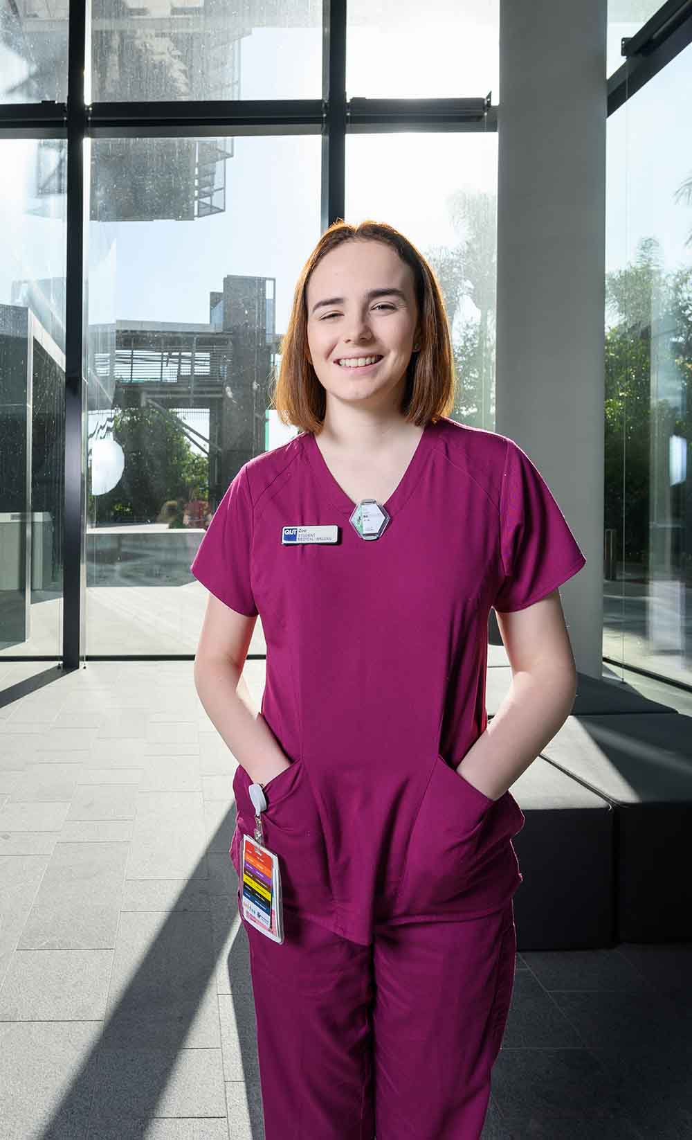 Zoe stands in a foyer, she's wearing her clinical scrubs and a name badge. Her access swipe cards are clipped to her pockets on an extendable cord..