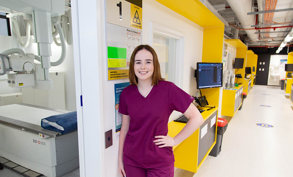 A female medical imaging student in scrubs stands in a hallway - next to hear is a treatment room with a bed.
