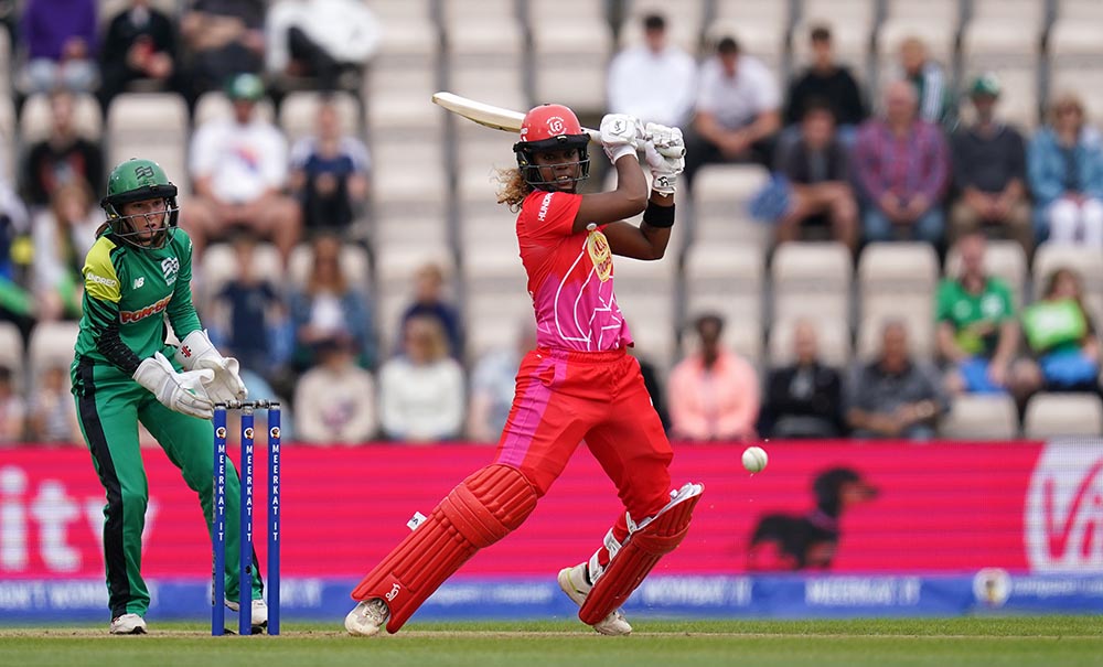 A female cricket batter dramatically swings at a cricket ball. She and the wicket keeper behind her are wearing full protective gear including helmets.