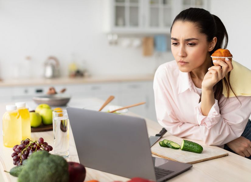 A woman sits at a table with an open laptop infront of her. She is frowning as she concentrates on the information around her is food and she's holding a muffin.