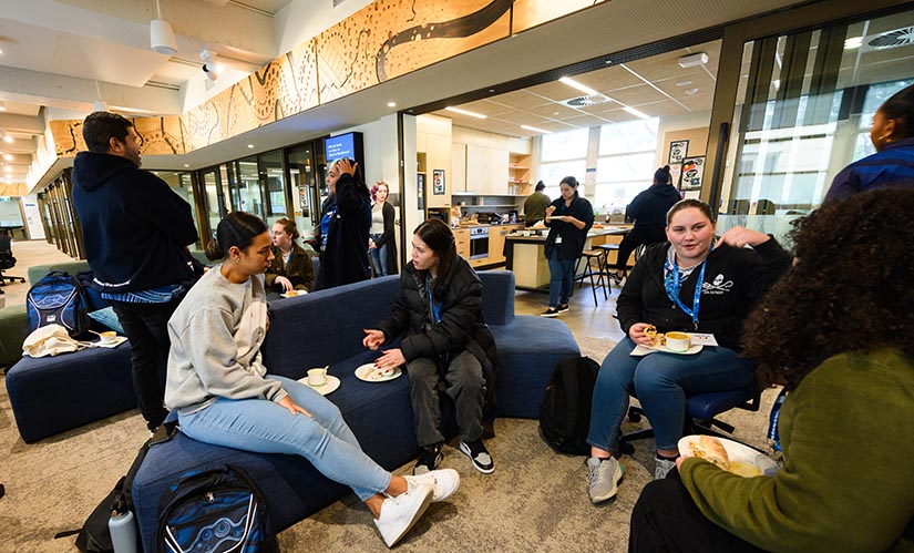 A group of students gather in modern, warmly lit facilities in the Oodgeroo Unit. They chat and laugh while sitting on comforatble couches and a woman in making tea in the kitchen facilities.