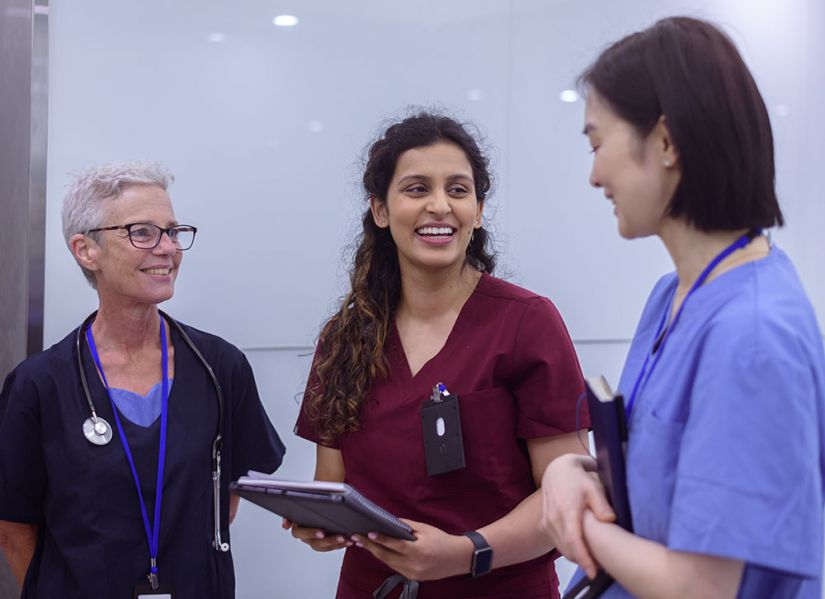 A woman in burgundy nursing scrubs smiles at one of her two supervisors in a hospital hallway.