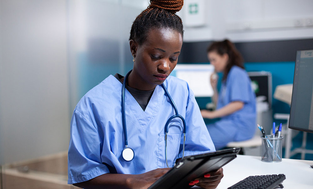 A nurse in scrubs with braids piled up on her head sits at a desk and reads through notes.