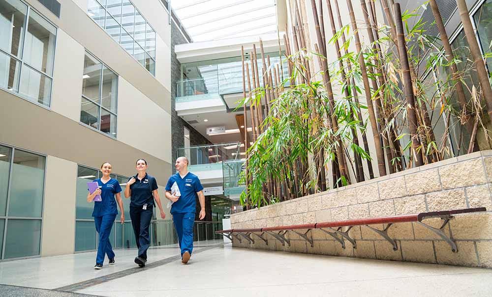 Three students carrying books and backpacks walk through a brightly-lit, high-ceilinged foyer in a QUT Health facility.