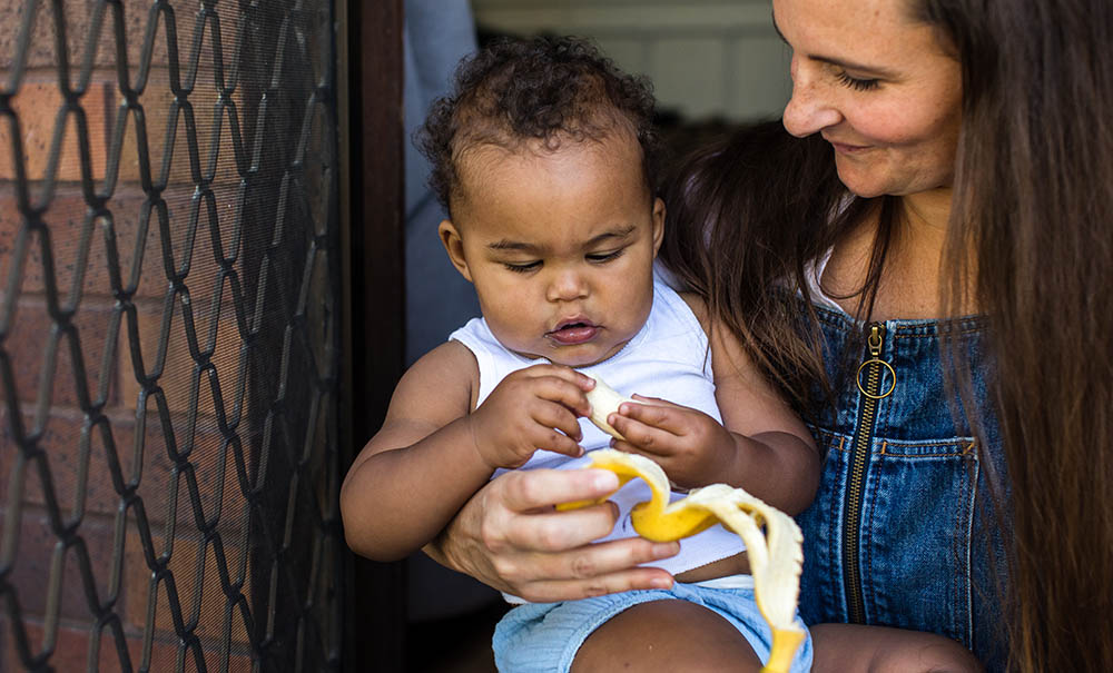 A mum sits with a toddler on her lap, he is eating a banana squishing it between his fingers.