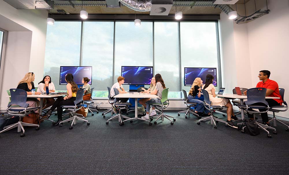 Students sit at group study tables in a large, modern room.
