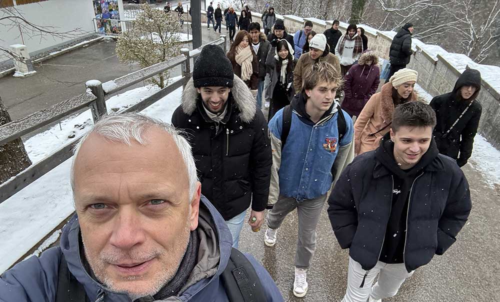 A male professor leads a group of laughing students as they walk along a snowy path.
