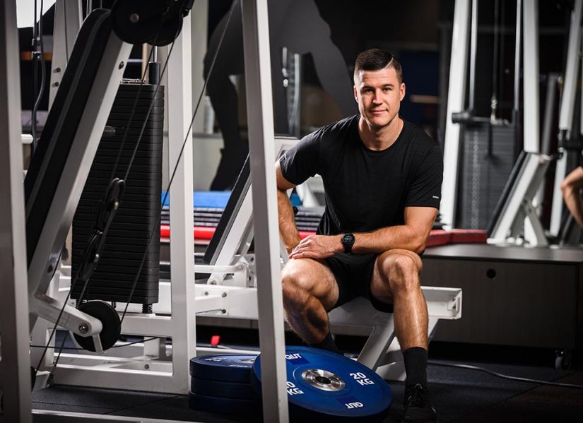 A clean cut, strong-looking guy wearing black, work out gear sitting on equipment in a gym.