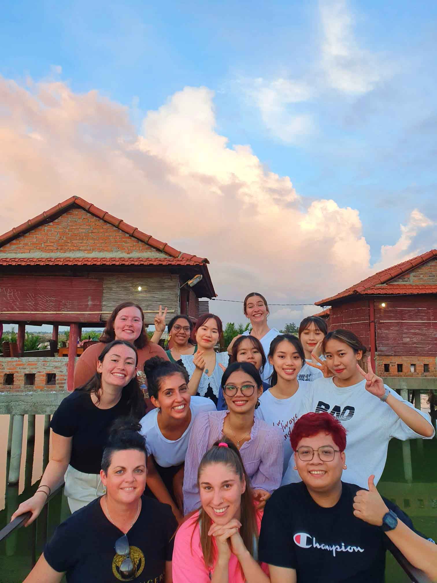 Happy students taking a selfie in warm afternoon light in front of wooden Vietnamese buildings.