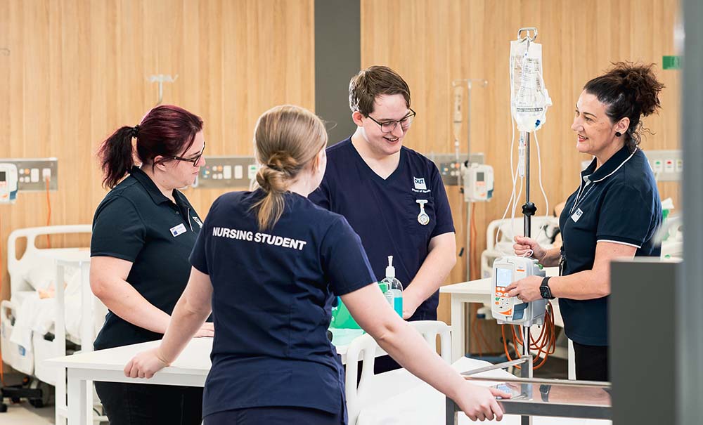 A teacher and three students stand next to a bed in a simulation laboratory that looks like a hospital ward.