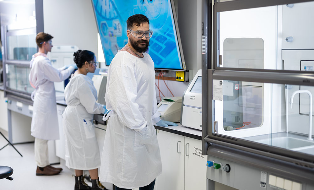 Three students in white lab coats and and protective glasses stand at a bench in a laboratory examining equipment.