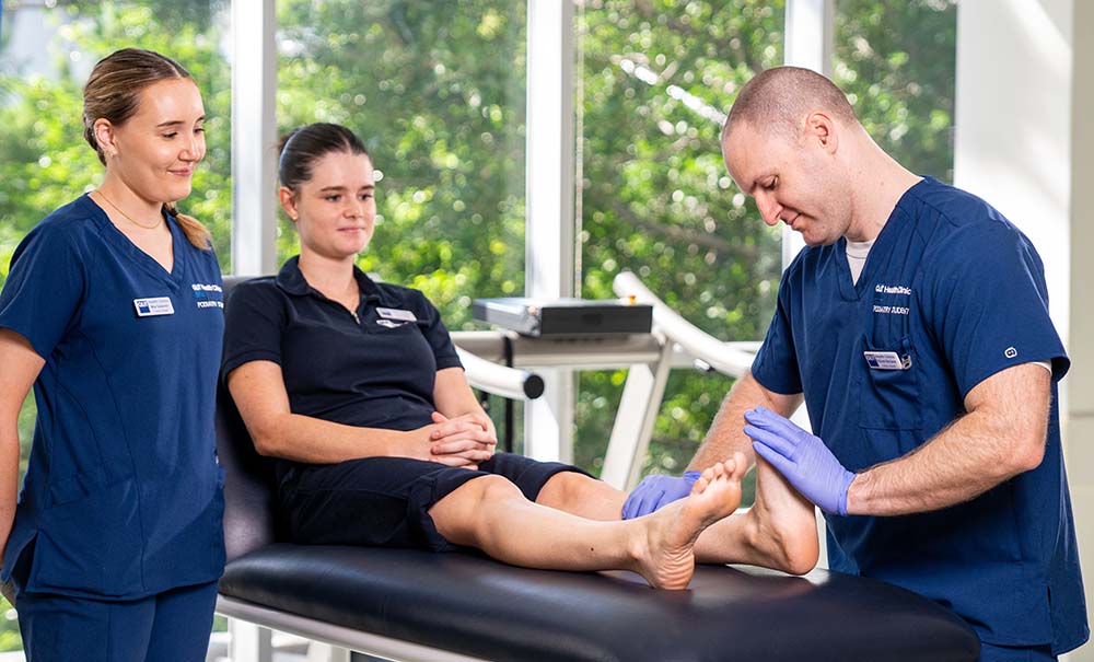 A female student is sitting barefoot on a bench. A male student, wearing gloves is testing the range of motion of her ankle.