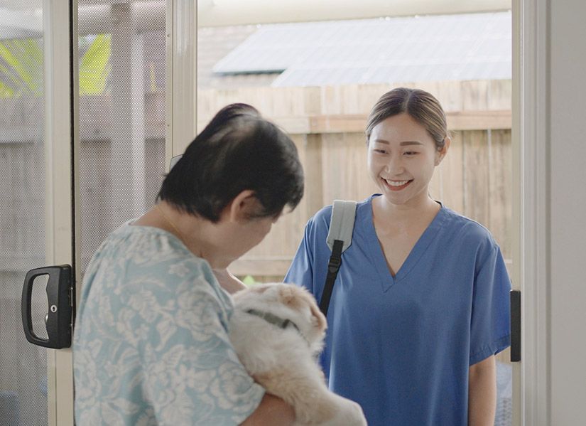 A female nurse in scrubs arrives as a home visit and is greeted by an elderly woman holding a small dog.