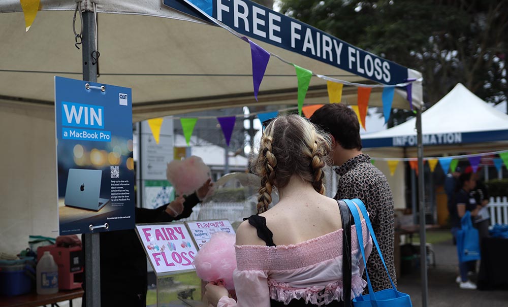 A girl with two thick braids wearing a pink corset standing infront of tent where free fairy floss is being handed out.