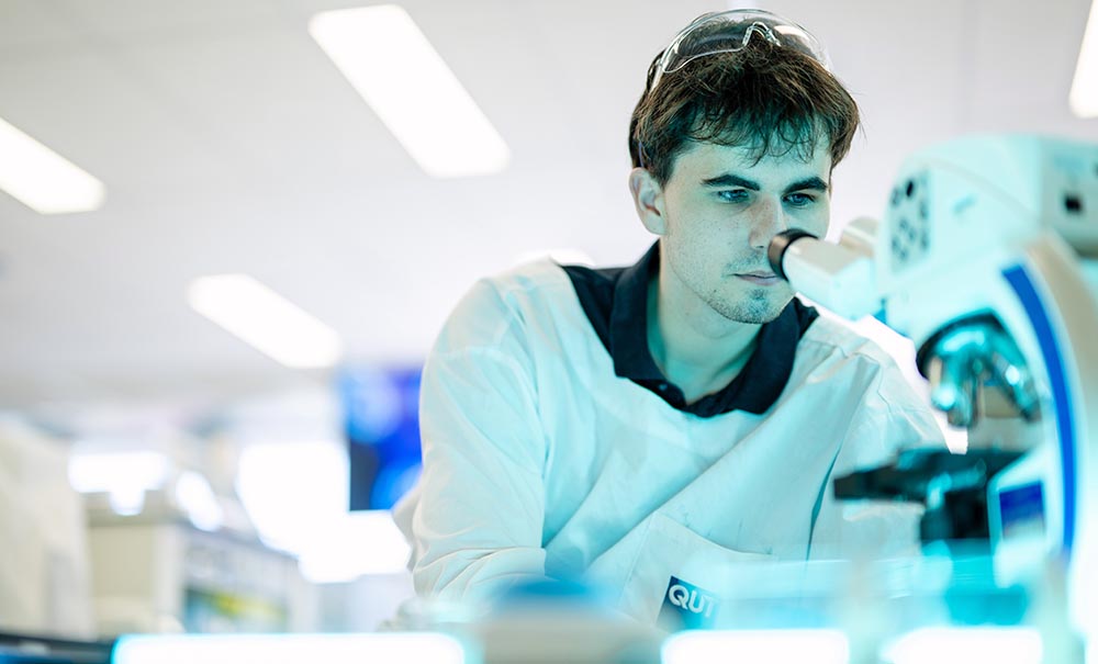 A male student in a laboratory looks down a microscope at a sample.