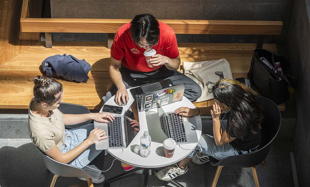 A group of students sit at a small table. Infront of them are laptops, note books, text books and coffee cups.